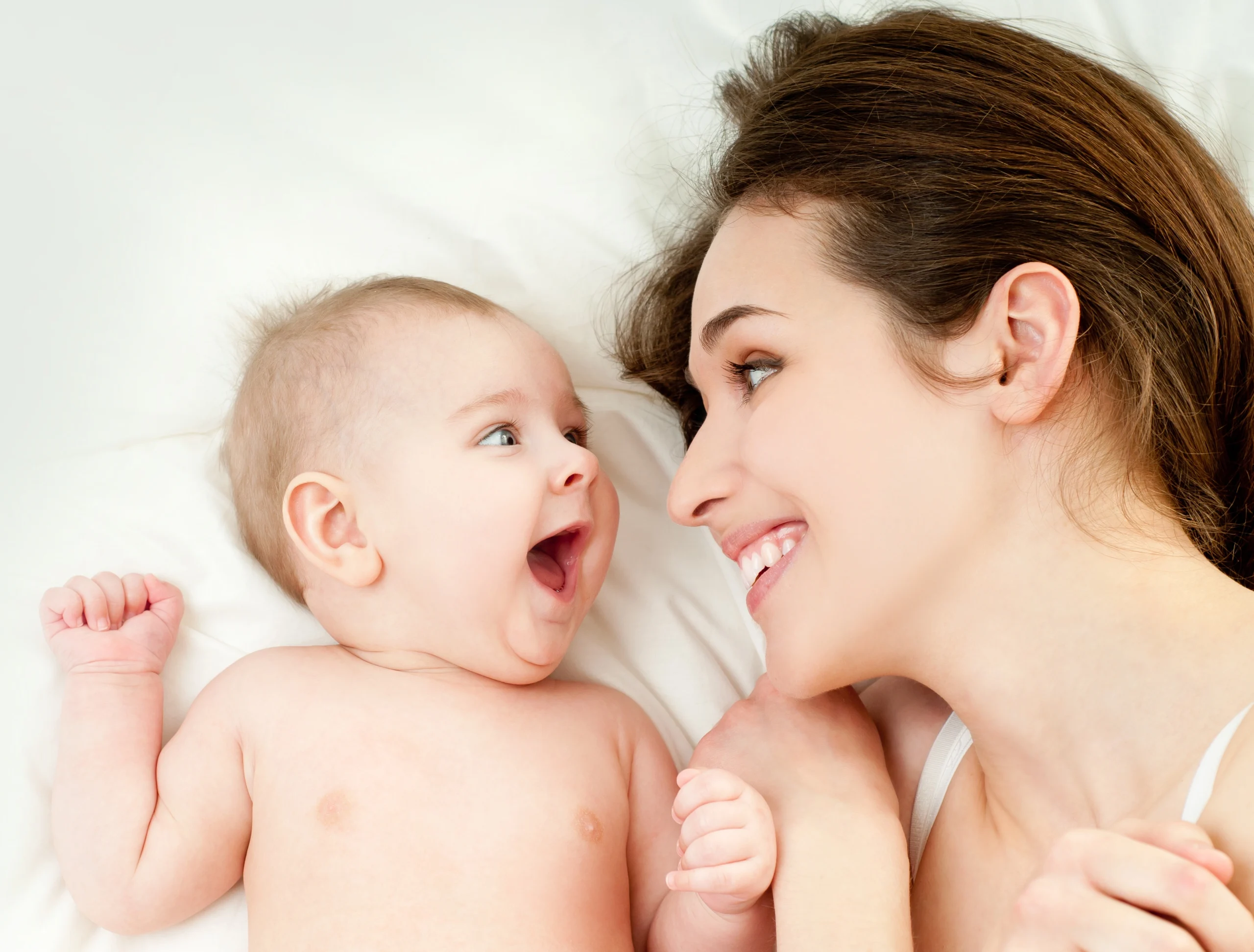 W jaki sposób mikrobiom matki wpływa na florę jelitową noworodków? Mama i niemowlę leżą na łóżku w białej pościeli i uśmiechają się do siebie.