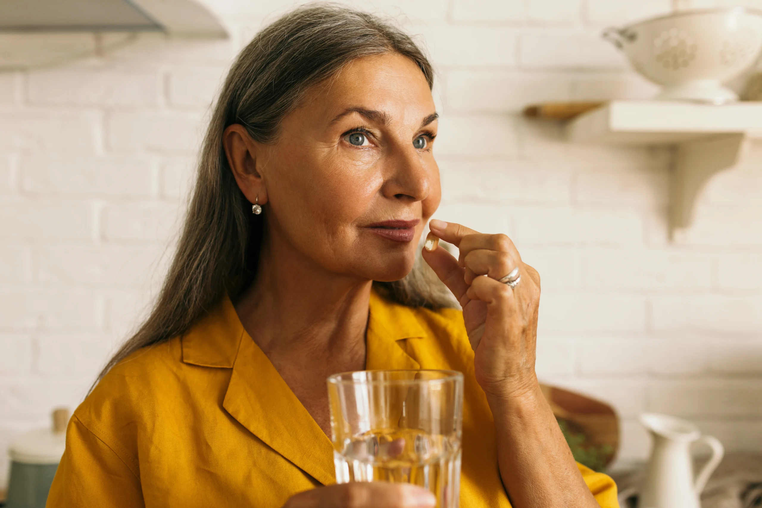Probiotyki dla seniorów – kiedy warto je stosować? Seniorka w żółtej bluzce przyjmuje lek/suplement/preparat probiotyczny.