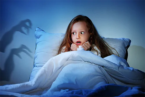 Co zrobić, gdy dziecko boi się ciemności? Dziewczynka skrywa się w łóżku pod kołdrą - boi się, że z ciemności wyjdą potwory - na ścianie widać cienie szponów.