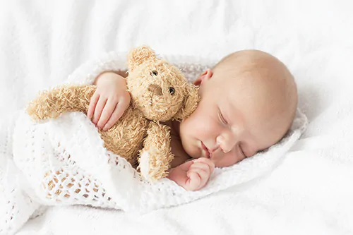 Jak długo powinno spać niemowlę? Słodki śpiący noworodek w zawiniątku przytula misia.