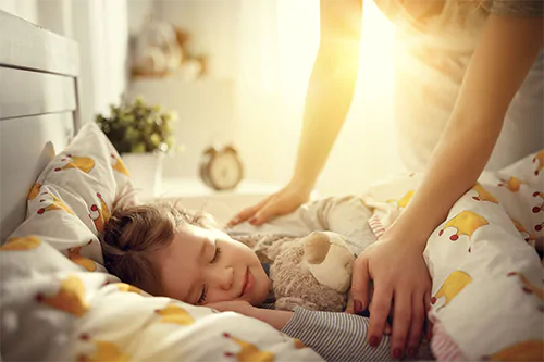 Jak nauczyć dziecko spania we własnym łóżku? Mama przykrywa kołdrą córeczkę śpiącą we własnym łóżeczku w pokoju dziecięcym.
