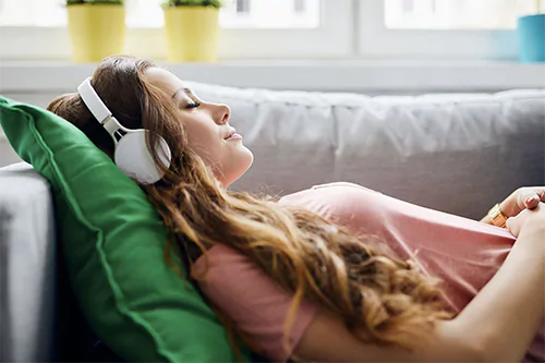 Jak radzić sobie ze stresem i napięciem? Kobieta ze słuchawkami na uszach leży na kanapie i słucha muzyki.
