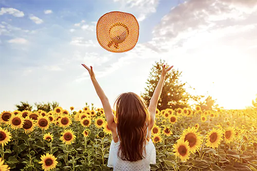 Jak wzmocnić odporność u dorosłych? Młoda kobieta stoi w polu słoneczników i podrzuca do góry słomkowy kapelusz.