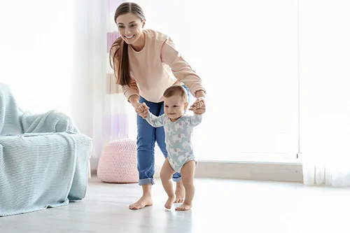 Nauka chodzenia - jak nauczyć dziecko chodzić? Mama prowadzi dziecko za rączki przez pokój.