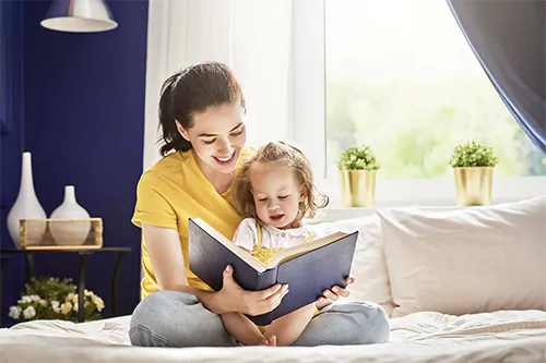 Nauka czytania - jak nauczyć dziecko czytać? Mama czyta z córeczką książkę, siedząc na łóżku w sypialni.