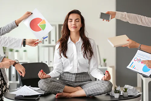 Objawy stresu - jak sobie z nimi radzić? Co na biegunkę ze stresu? Businesswoman siedzi na biurku w pracy i medytuje. Wokół niej wyciągnięte ręce z dokumentami.