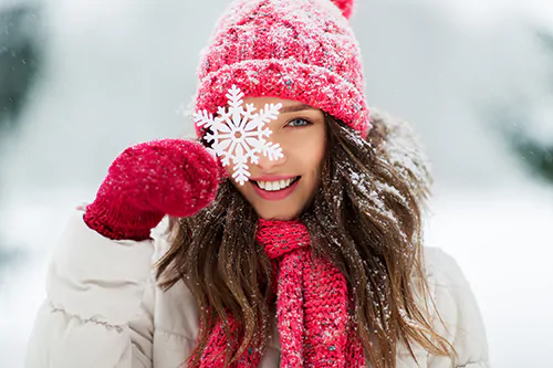 Jak wzmocnić odporność zimą? Młoda kobieta w czerwonej czapce, stojąca na śniegu, przykłada ozdobę w postaci płatka śniegu do twarzy.