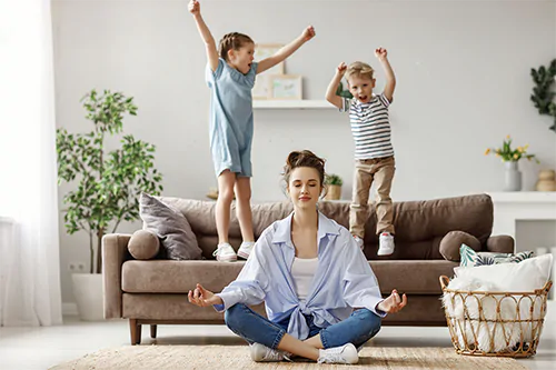 Równowaga między pracą a życiem osobistym - jak znaleźć work-life balance? Kobieta medytuje w salonie, podczas gdy jej dzieci szaleją na kanapie.