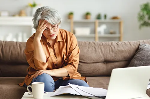 Skutki stresu - czy długotrwały stres jest groźny dla zdrowia? Seniorka w rudopomarańczowej koszuli i okularach siedzi zmartwiona na kanapie w salonie.