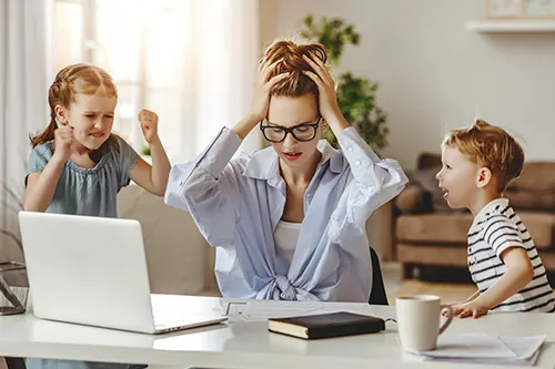 Stres w pracy - jak sobie z nim radzić i czym grozi długotrwałe napięcie? Młoda sfrustrowana mama pracuje zdalnie w domu przy laptopie, ale przeszkadzają jej w tym małe dzieci, biegające wokół niej.