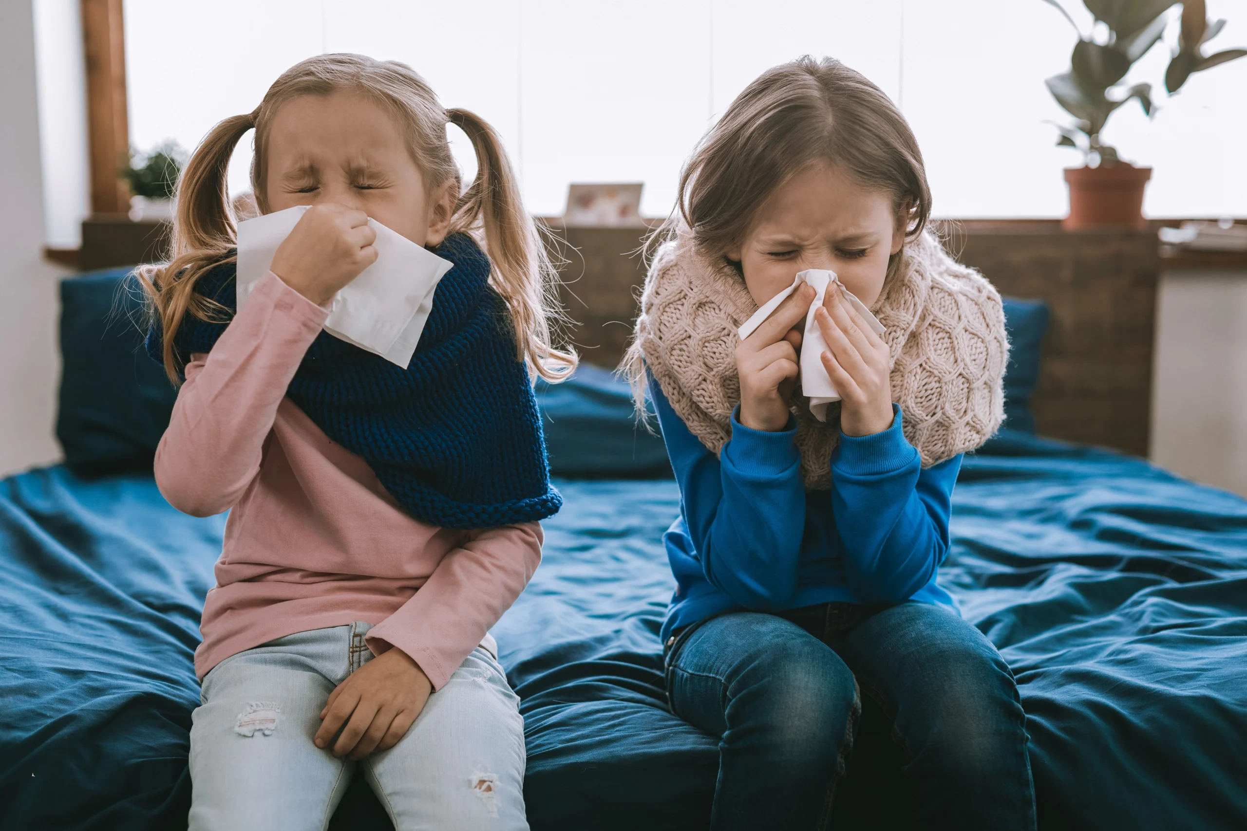 Jak podnieść obniżoną odporność u dzieci? Jakie są sposoby na obniżoną odporność? Dwie zakatarzone dziewczynki siedzą na łóżku w pokoju z chusteczkami przy nosach.