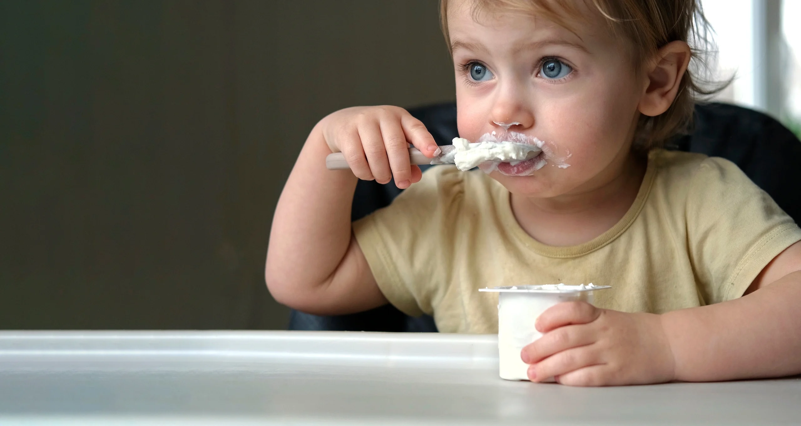 Sprawdź, jakie probiotyki dla dzieci wybrać i kiedy je stosować. Małe dziecko siedzi przy stole kuchennym i je serek lub jogurt łyżeczką.