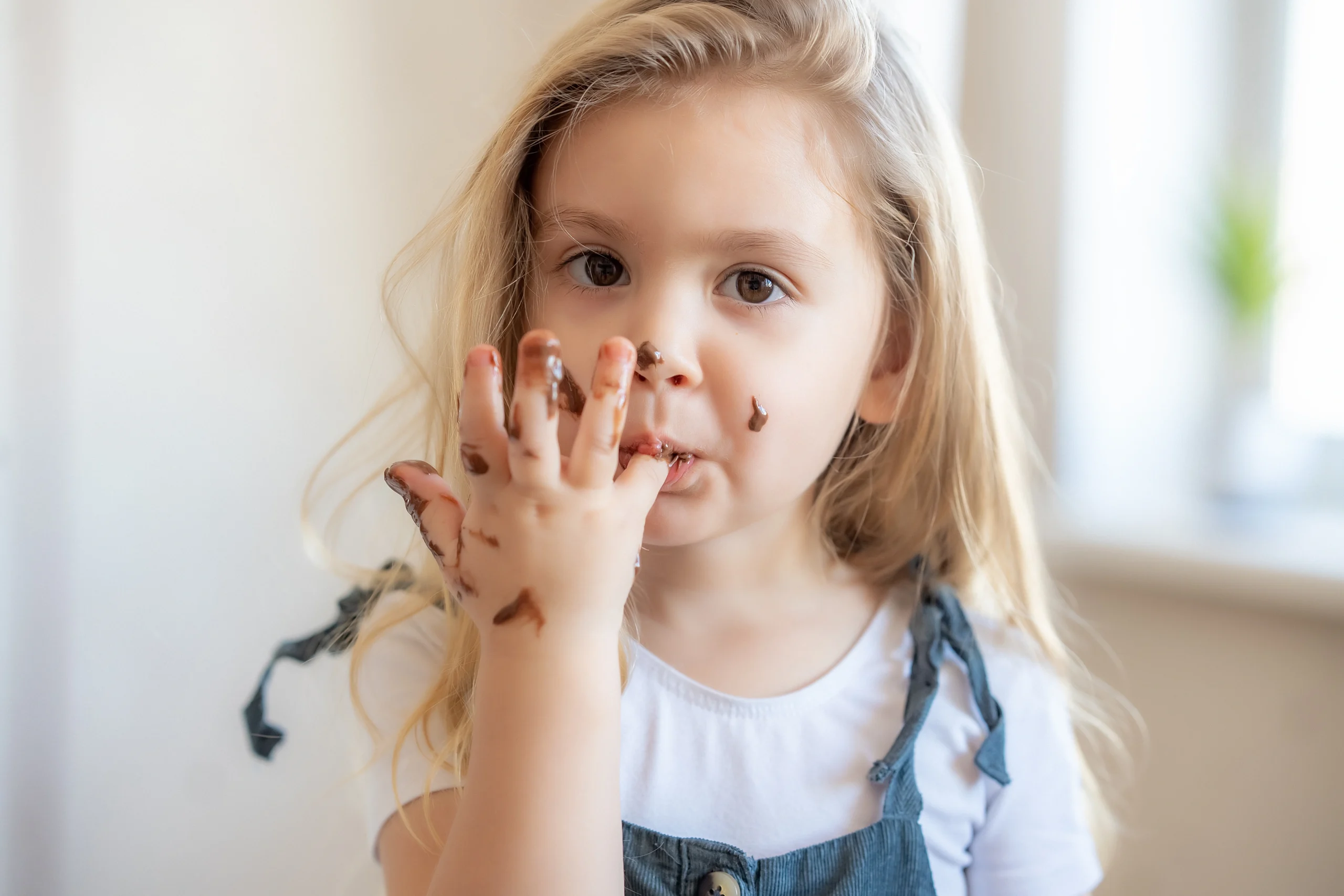 Czekoladki probiotyczne dla dzieci - jak i kiedy je stosować? Mała dziewczynka w długich blond włosach oblizuje palce po zjedzeniu czekolady.