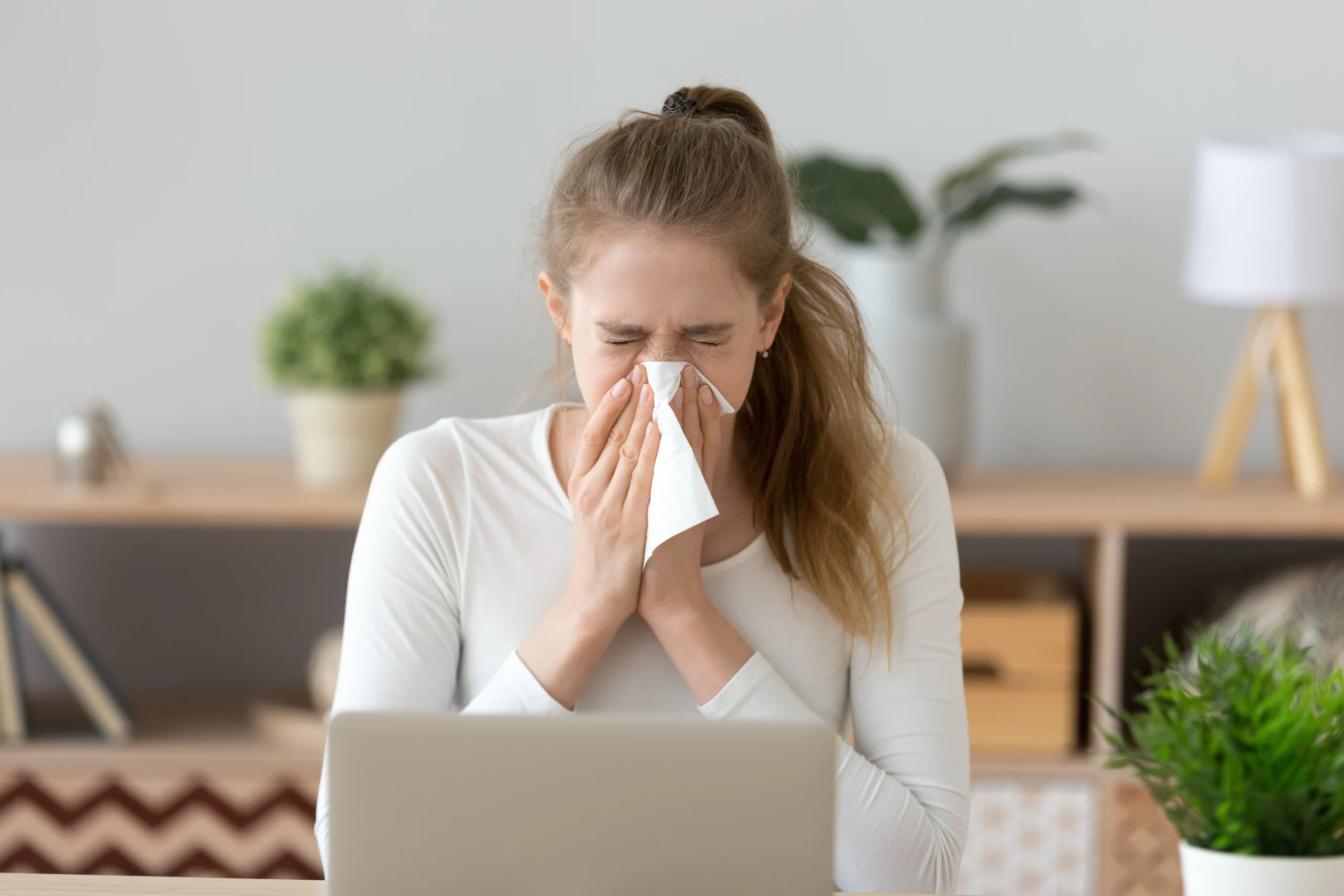 Jakie są czynniki obniżające odporność u dorosłych? Kobieta z katarem, siedząca przy laptopie, wydmuchuje nos w chusteczkę.