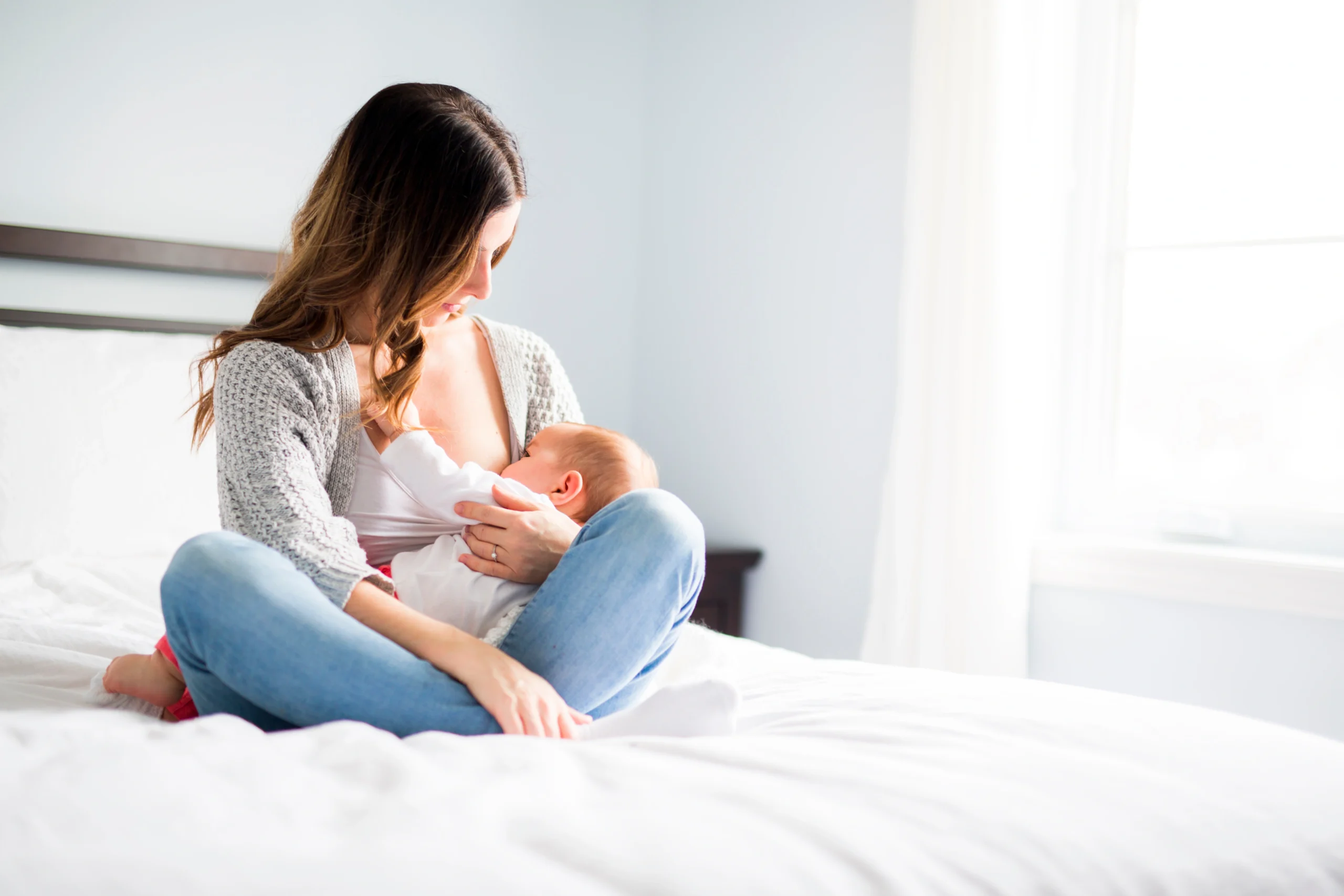Jakie znaczenie ma mikrobiota jelitowa w pierwszych tygodniach życia dziecka? Mama karmi noworodka piersią, siedząc na łóżku w sypialni.