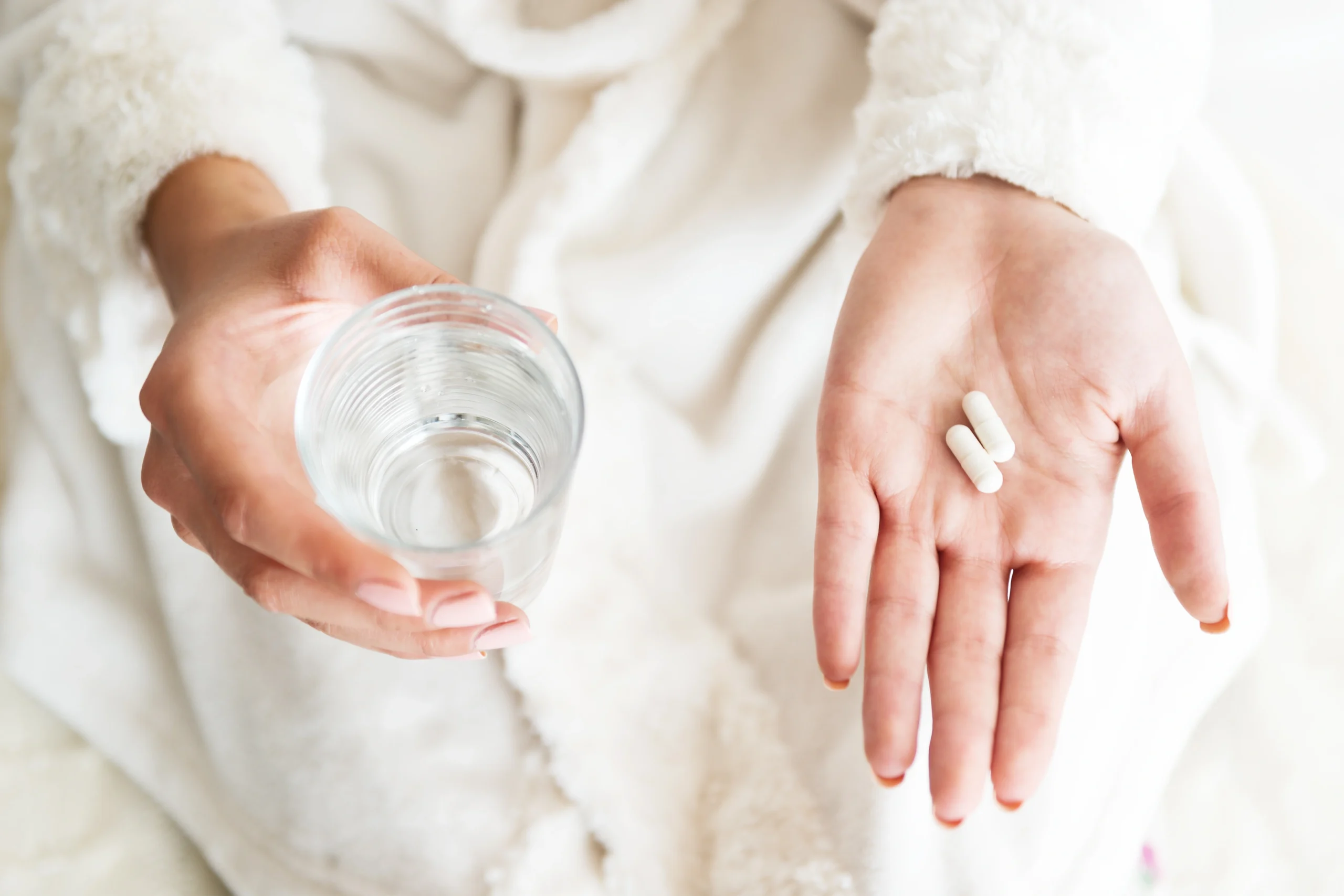 Synbiotyk - co to jest i czym się wyróżnia? Czym są synbiotyki? Zbliżenie na kobiece dłonie trzymające 2 kapsułki leku oraz szklankę z wodą.