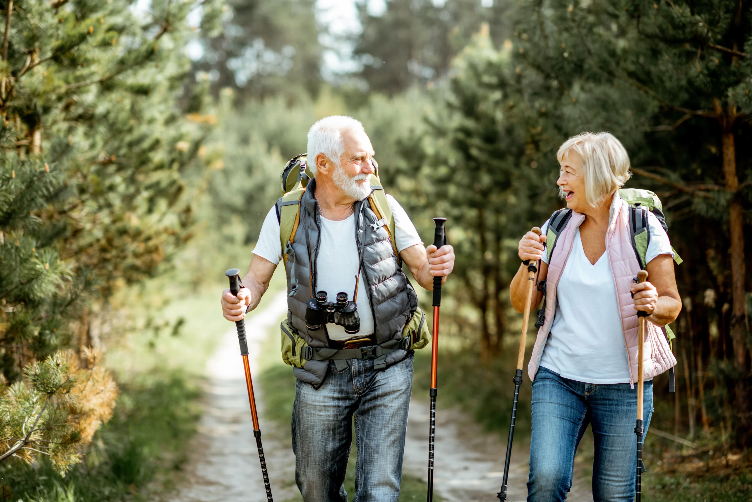 Aktywny senior na emeryturze - dlaczego nie warto siedzieć na kanapie? Senior i seniorka z plecakami uprawiają nordic walking w lesie.