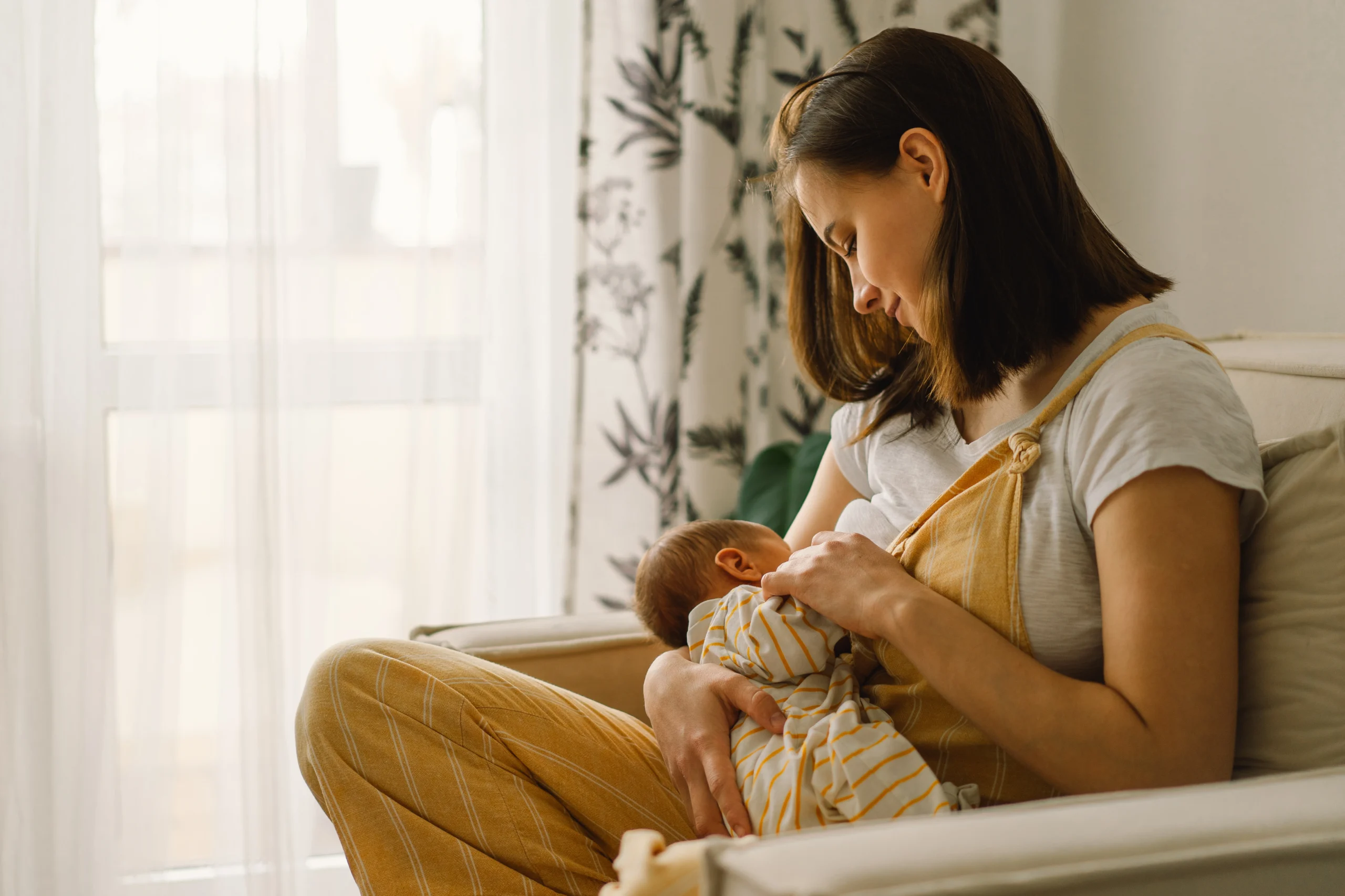 Jak karmienie piersią wpływa na mikrobiom u noworodków i niemowląt? Mama karmi noworodka piersią, siedząc przy oknie w fotelu.