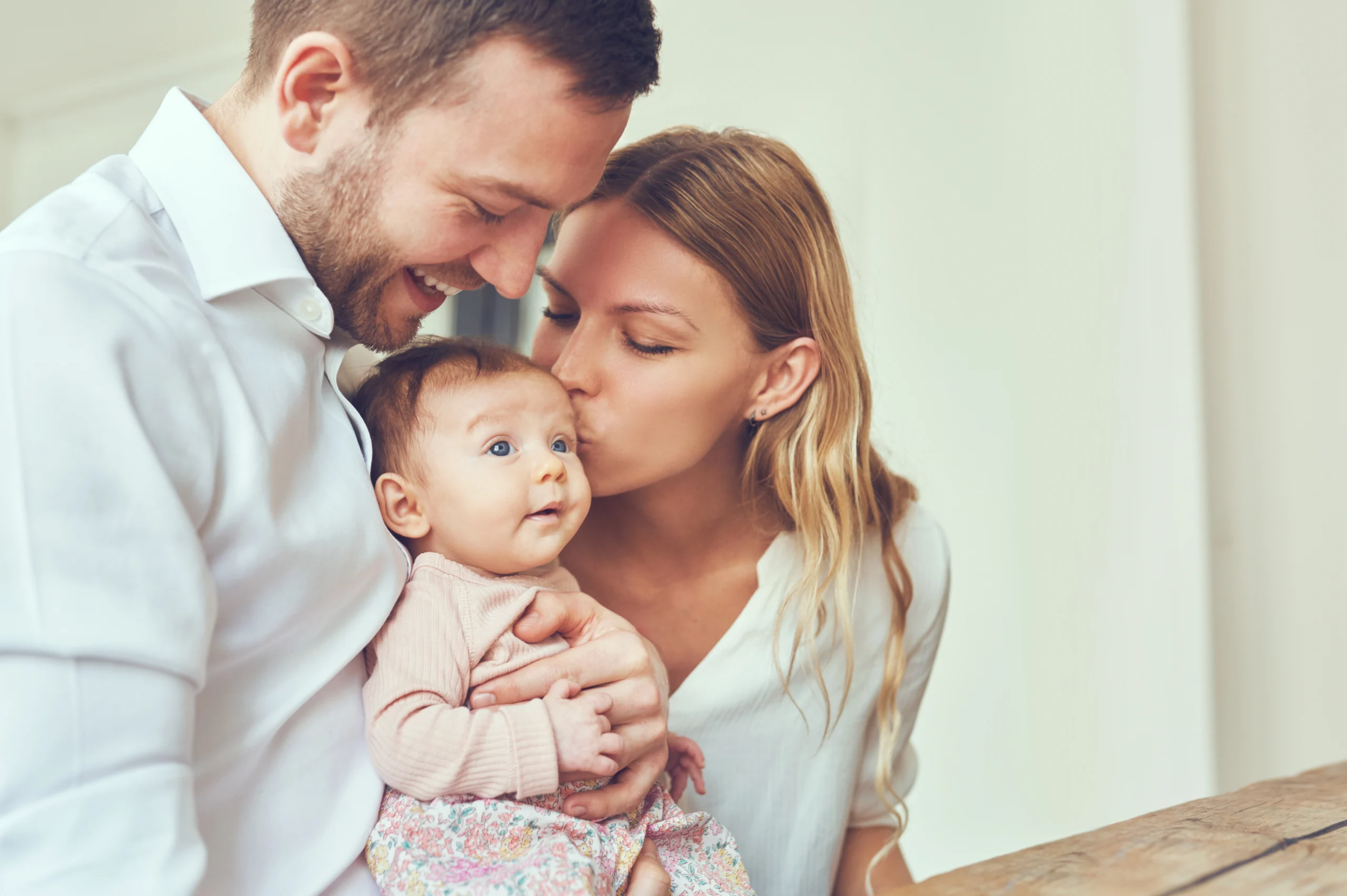 Czy mikrobiom niemowlaka i dorosłego jest taki sam? Jakie są pomiędzy nimi różnice? Czy mikrobiom można kształtować na przestrzeni lat? Mama i tata trzymają na rękach niemowlaka., a mama całuje dziecko w główkę.