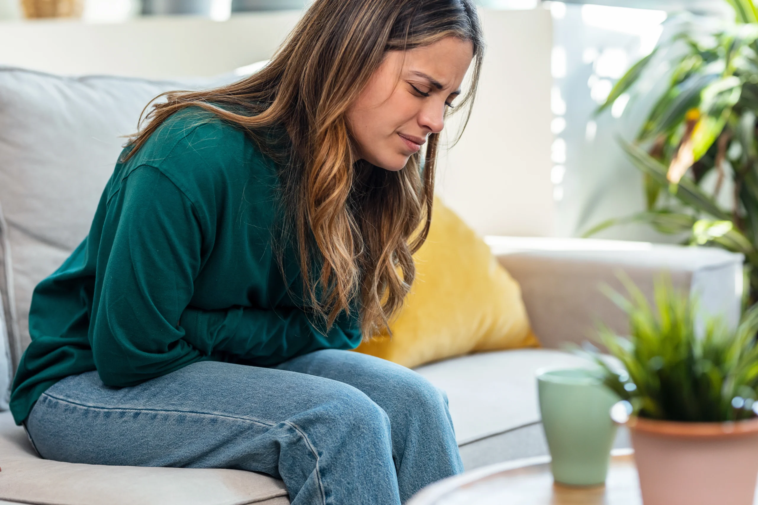 Czy osłabienie po antybiotyku jest normalne i jak sobie pomóc? Na jakie objawy i skutki uboczne po antybiotyku zwrócić uwagę? Kobieta w zielonej bluzie i jeansach siedzi na kanapie w salonie i trzyma się za bolący brzuch.