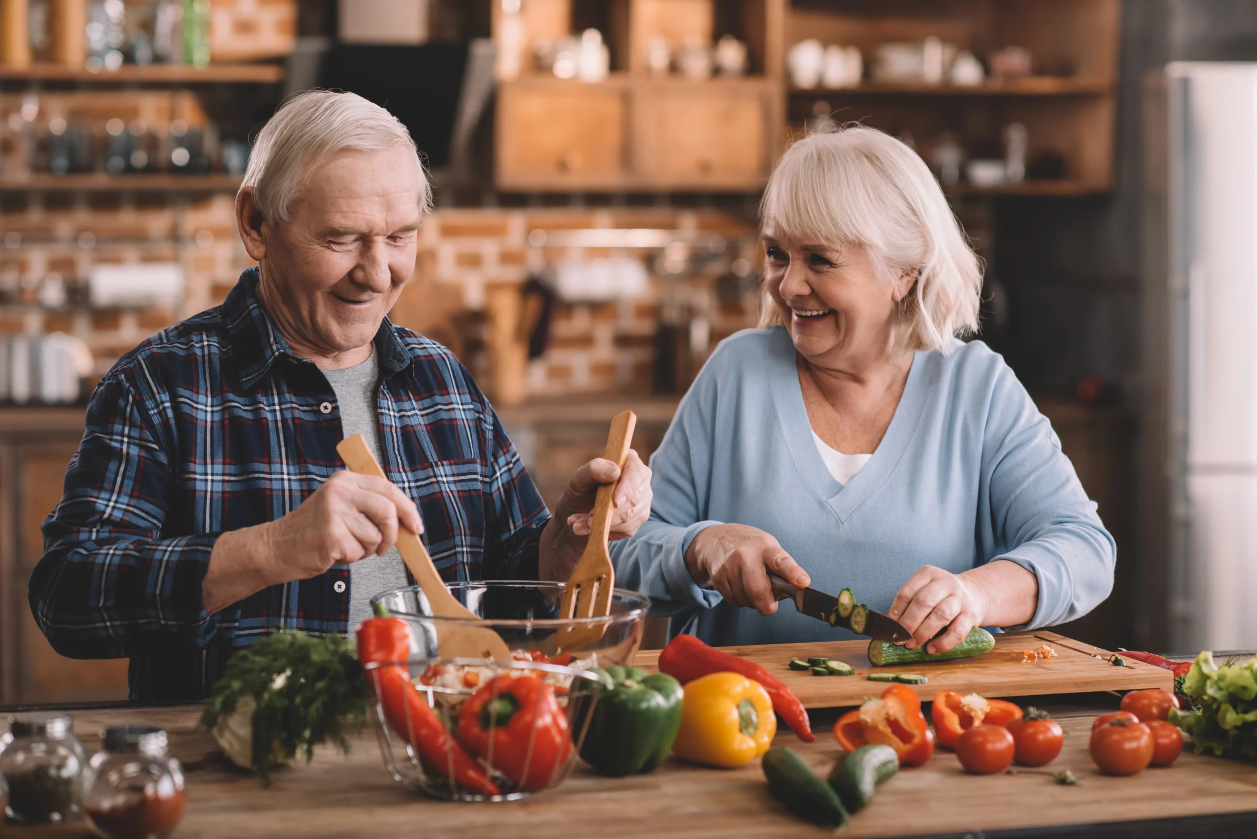 Para uśmiechniętych seniorów przygotowuje w kuchni wspólny posiłek na bazie kolorowych warzyw i innych zdrowych produktów, będących elementem zdrowej diety dla osób po 60, 70 i 80 roku życia.